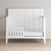 The Child Craft Atwood 4-in-1 Convertible Crib in Matte White features a chevron-patterned back panel, partial side rail, and mattress, set on a light wood floor against a plain beige wall.