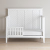 The Child Craft Toddler Guard Rail in Matte White features sturdy wood construction and a partial guard rail with a herringbone back panel, shown against a light wall on a hardwood floor.