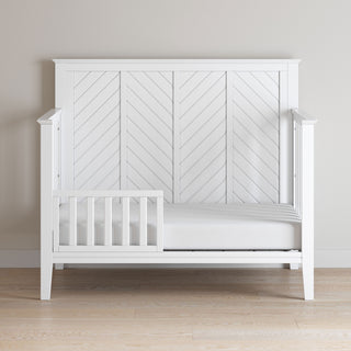 The Child Craft Toddler Guard Rail in Matte White features sturdy wood construction and a partial guard rail with a herringbone back panel, shown against a light wall on a hardwood floor.