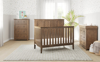 A bright nursery with a wooden crib, Child Craft Atwood 3-Drawer Dresser in Cocoa Bean, changing table, gray rug, pastel wallpaper, a potted plant, and a window with sheer curtains letting in natural light.