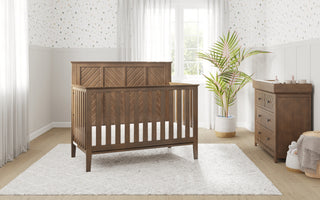 A wooden crib sits on a white rug in a bright nursery with light walls, a Child Craft Atwood 3-Drawer Dresser in Cocoa Bean, a large potted plant, and a window with sheer curtains letting in sunlight.