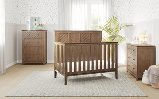 A modern nursery with a wooden crib, Child Craft Atwood 3-Drawer Dresser in Cocoa Bean, matching chest, gray rug, plant, soft lighting, white walls with decorative wallpaper, and a large window with sheer curtains.