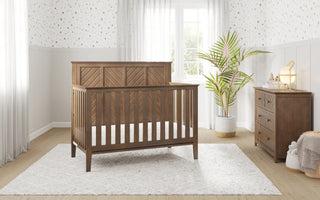 The Child Craft Atwood 4-in-1 Convertible Crib in Cocoa Bean stands on a white rug in a bright nursery with white walls, a potted plant, and a wooden dresser, bathed in natural light from sheer curtains.
