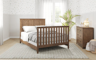 A cozy bedroom featuring the Child Craft Atwood 4-in-1 Convertible Crib in Cocoa Bean, with white bedding, a light gray rug, matching wooden dresser, potted plant by the window, white paneled walls and patterned wallpaper filled with natural light.