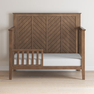 A Child Craft F09501 Toddler Guard Rail in Cocoa Bean is installed on a wooden toddler bed with chevron headboard, partial rails, and white mattress, positioned on a light wood floor against a pale wall for added safety.