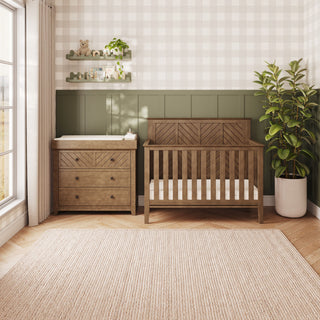 An Atwood 4-in-1 Convertible Crib in cocoa bean and a matching three-drawer changing table with a pad, set in a nursery with a beige rug and green-and-white paneled walls.