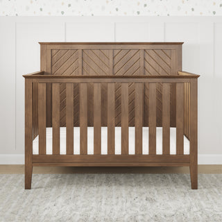 The Child Craft Atwood 4-in-1 Convertible Crib in Cocoa Bean features a geometric headboard design and vertical slats, shown here on a light rug against a white paneled wall with patterned wallpaper above.