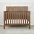 The Child Craft Atwood 4-in-1 Convertible Crib in Cocoa Bean features a geometric headboard design and vertical slats, shown here on a light rug against a white paneled wall with patterned wallpaper above.