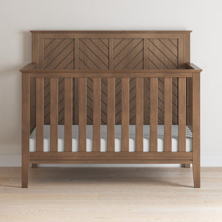 The Child Craft Atwood 4-in-1 Convertible Crib in Cocoa Bean features a chevron-patterned headboard and vertical slats, shown on a light wood floor against a plain white wall.