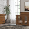 A nursery with a wooden crib, matching dresser featuring the Child Craft Universal Dressing Kit in Coach Cherry, a potted plant, large window with white curtains, framed wall art, and a gray area rug.