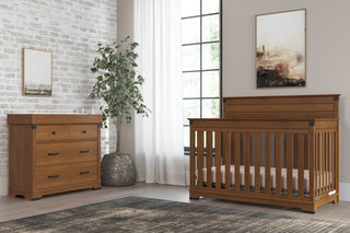 A nursery with a wooden crib, matching dresser featuring the Child Craft Universal Dressing Kit in Coach Cherry, a potted plant, large window with white curtains, framed wall art, and a gray area rug.
