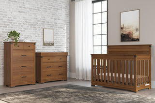 A modern nursery features wooden furniture, including a crib and a Child Craft Universal Dressing Kit in Coach Cherry, with natural light from sheer-curtained windows. A plant and framed art add cozy décor against the white brick wall.