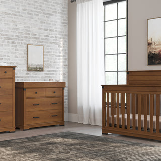 A nursery room features the Child Craft Redmond 4 Drawer Chest in Coach Cherry, paired with a wooden crib and dresser against white brick and beige walls with framed art. A plant decorates the chest as sunlight filters through white curtains.