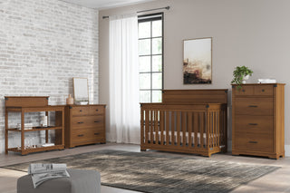 A modern nursery with wooden furniture, including a crib, a tall dresser, a changing table, and the Child Craft Redmond 3 Drawer Dresser in Coach Cherry. Features include a gray rug, light walls, brick accent wall, window with white curtains, and a plant.