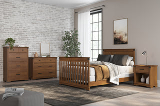 A modern bedroom with a wooden bed, nightstand, dresser, and the Child Craft Redmond 4-in-1 Convertible Crib in Coach Cherry. Neutral walls, sheer curtains on a large window, gray rug, plant, and minimal wall art complete the space.