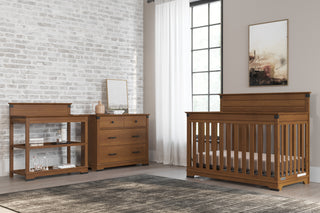 A sunlit, modern nursery features a wooden crib, the Child Craft Redmond 3 Drawer Dresser in Coach Cherry, and a changing table against a brick accent wall, plus sheer curtains, neutral walls, and framed abstract art.