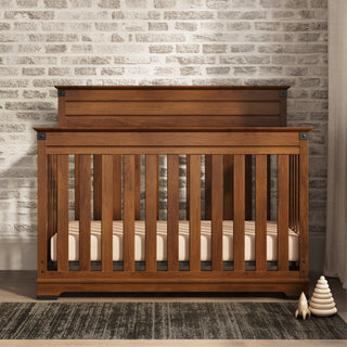 A Child Craft Redmond 4-in-1 Convertible Crib in Coach Cherry with vertical slats stands against a white brick wall. Nearby are a striped rug, a wooden stacking toy and cone, and pale curtains hanging to the right.
