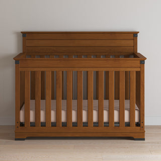 A Child Craft Redmond 4-in-1 Convertible Crib in Coach Cherry with vertical slats and a high backrest stands against a light wall on a wooden floor, featuring a light-colored mattress inside.