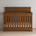 A Child Craft Redmond 4-in-1 Convertible Crib in Coach Cherry with vertical slats and a high backrest stands against a light wall on a wooden floor, featuring a light-colored mattress inside.
