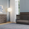 A modern nursery with blue walls, a wooden crib, a Child Craft Redmond 3 Drawer Dresser in Dapper Gray, a floor lamp, gray rug, sheer curtains, and abstract art. Sunlight from a large window brightens the cozy space.