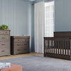 A nursery with a dark wooden crib, matching dresser, and the Child Craft Redmond 4 Drawer Chest in Dapper Gray stands against pale blue paneled walls, with a large window, white curtains, a rug, and an orange ottoman on the floor.