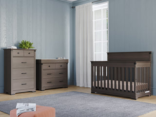 A nursery with light blue walls, a tall dresser, a changing table, a Child Craft Redmond 4-in-1 Convertible Crib in Dapper Gray, a gray rug, and a large window with white curtains letting in natural light.
