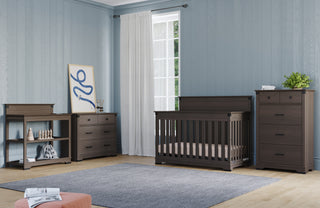 A nursery with light blue walls, a window with white curtains, a brown crib, a Child Craft Redmond 3 Drawer Dresser in Dapper Gray, matching dressers, a bookshelf with toys, a gray rug, and wall art resting on a dresser.