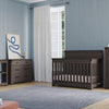 A cozy nursery with light blue walls, a window with white curtains, and dark wood furniture, featuring the Child Craft Redmond 4 Drawer Chest in Dapper Gray, plus a crib, dresser, toys on shelves, a rug, and framed artwork.
