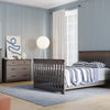 A modern bedroom with light blue walls, gray wooden furniture, a bed with striped bedding, a Child Craft Redmond 3 Drawer Dresser in Dapper Gray, a wide dresser with art and a plant on top, a nightstand, round floor lamp, and a rug on light wood floors.