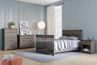 A bedroom with light blue walls and wooden floors features a gray bed, a dresser, a chest of drawers, a modern lamp, a rug, minimal decor including plants and an abstract print, plus the Child Craft Redmond Nightstand in Dapper Gray.