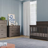 A nursery features gray wooden furniture: a crib, Child Craft Redmond 3 Drawer Dresser in Dapper Gray, and a tall chest. A modern print, plants, and vases top the dresser. Blue walls, a large window with sheer curtains, and a gray rug complete the look.