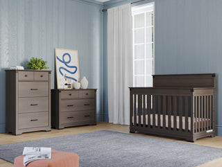 A modern nursery features gray wood furniture: a crib, a dresser with decor, and the Child Craft Redmond 4 Drawer Chest in Dapper Gray. Light blue walls, a gray rug, and a large window with white curtains brighten the room.