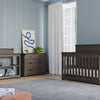A modern nursery features gray wooden furniture, including a crib, Child Craft Redmond 3 Drawer Dresser in Dapper Gray, and changing table. Light blue walls, a large window with white curtains, and a gray rug complete the room’s look.