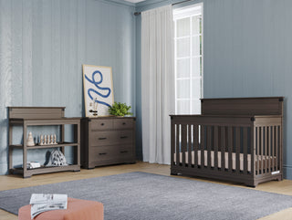 A modern nursery features gray wooden furniture, including a crib, Child Craft Redmond 3 Drawer Dresser in Dapper Gray, and changing table. Light blue walls, a large window with white curtains, and a gray rug complete the room’s look.