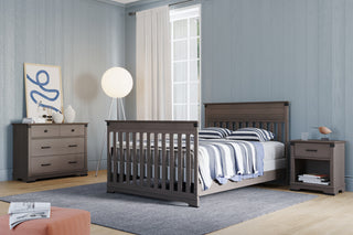 A modern bedroom features gray wood furniture, including a crib-style bed, nightstand, and the Child Craft Redmond 3 Drawer Dresser in Dapper Gray. Striped bedding, light curtains, a globe floor lamp, and abstract art complete the space.
