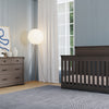 A modern nursery with light blue paneled walls, a gray crib, a Child Craft Redmond 3 Drawer Dresser in Dapper Gray with decor, a large round floor lamp, white curtains, and a textured gray rug on wooden floors.