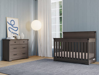 A modern nursery with light blue paneled walls, a gray crib, a Child Craft Redmond 3 Drawer Dresser in Dapper Gray with decor, a large round floor lamp, white curtains, and a textured gray rug on wooden floors.