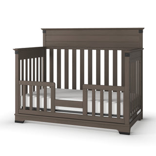 The Child Craft F09514 Toddler Guard Rail in Dapper Grey is shown installed on a brown wooden crib in toddler bed mode, featuring slatted sides, back, and a visible white mattress.