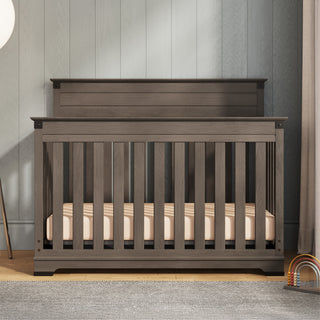 The Child Craft Redmond 4-in-1 Convertible Crib in Dapper Gray stands on a wooden floor by a paneled wall, with vertical slats. A rainbow toy is on the right, and a gray rug is partly visible in front of this stylish crib.