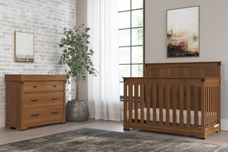 A cozy nursery features a wooden crib and the Child Craft Redmond 3 Drawer Dresser in Coach Cherry, a potted plant, large window with white curtains, framed art on the walls, and a gray rug on a wood floor.
