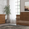 A cozy nursery with a wooden crib and matching dresser topped with the Child Craft F01792 Universal Dressing Kit in Coach Cherry, a potted plant, abstract art, sheer-curtained window for natural light, gray rug, and light floor.