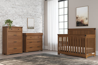A nursery with wooden furniture, including a crib and the Child Craft Redmond 3 Drawer Dresser in Coach Cherry, features light walls, a large window with white curtains, a gray rug, and framed art on the walls.