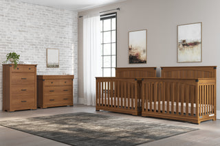 A modern nursery with two Child Craft Redmond Full Panel 4-in-1 Convertible Cribs in Coach Cherry, a dresser, and changing table by a brick accent wall. Features a large window with sheer curtains, abstract art, and a gray area rug.