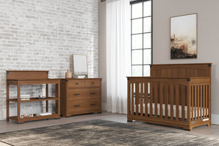 A cozy nursery showcases the Child Craft Redmond Full Panel 4-in-1 Convertible Crib in Coach Cherry, paired with matching dresser and changing table by a brick wall, with a rug underfoot and framed art on a beige wall.