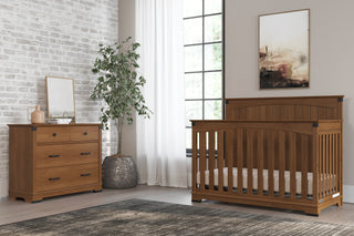 A nursery featuring the Child Craft Redmond Full Panel 4-in-1 Convertible Crib in Coach Cherry with a matching dresser, potted plant, rug, and neutral decor. A large window with white curtains fills the space with natural light.