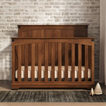 The Child Craft Redmond Full Panel 4-in-1 Convertible Crib in Coach Cherry stands on a dark rug before a white brick wall, with a stackable toy and small ornament nearby, creating a cozy nursery as light streams in from the right.