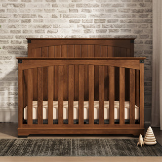 The Child Craft Redmond Full Panel 4-in-1 Convertible Crib in Coach Cherry stands on a dark rug before a white brick wall, with a stackable toy and small ornament nearby, creating a cozy nursery as light streams in from the right.