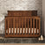 The Child Craft Redmond Full Panel 4-in-1 Convertible Crib in Coach Cherry stands on a dark rug before a white brick wall, with a stackable toy and small ornament nearby, creating a cozy nursery as light streams in from the right.