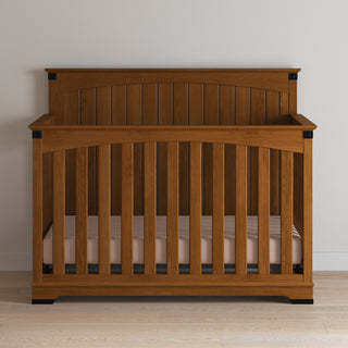 The Child Craft Redmond Full Panel 4-in-1 Convertible Crib in Coach Cherry features vertical slats and a curved headboard, set against a light wall and floor. A beige mattress sheet completes this stylish nursery furniture.