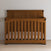 The Child Craft Redmond Full Panel 4-in-1 Convertible Crib in Coach Cherry features vertical slats and a curved headboard, set against a light wall and floor. A beige mattress sheet completes this stylish nursery furniture.